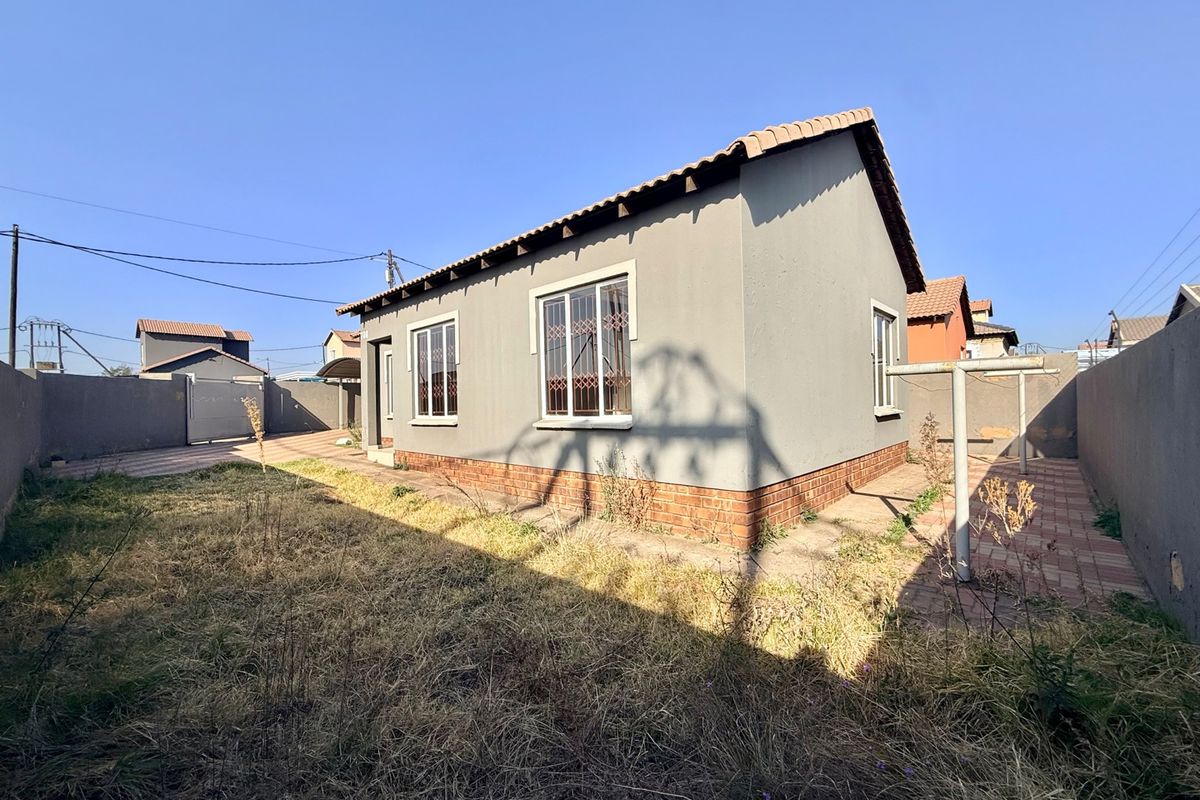 Rear view of a detached house featuring a tiled roof, brick plinth, security bars on windows, and a paved area with an overgrown garden.