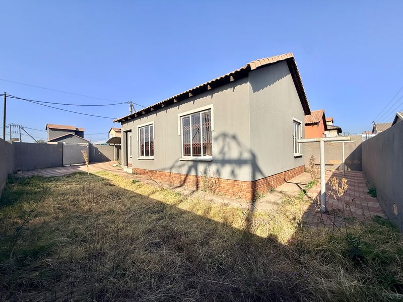 Rear view of a detached house featuring a tiled roof, brick plinth, security bars on windows, and a paved area with an overgrown garden.