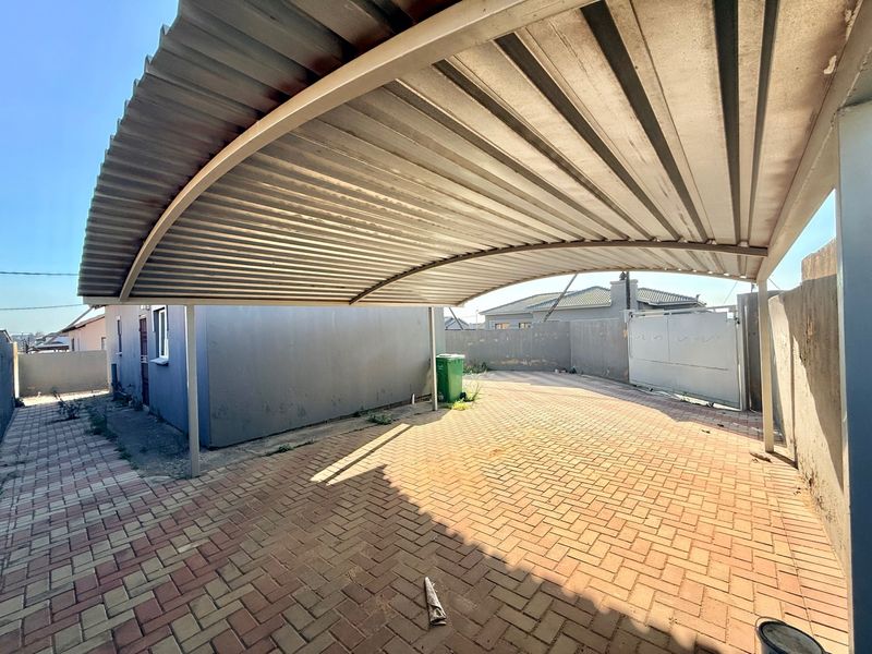 A paved driveway featuring a curved corrugated metal carport structure, enclosed by boundary walls and a sliding gate.