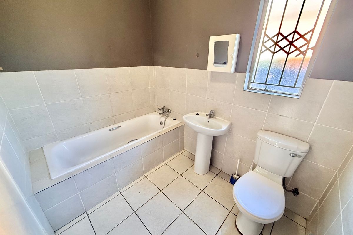 A functional bathroom featuring a bathtub, pedestal basin, and toilet with neutral wall and floor tiling and security bars on the window.