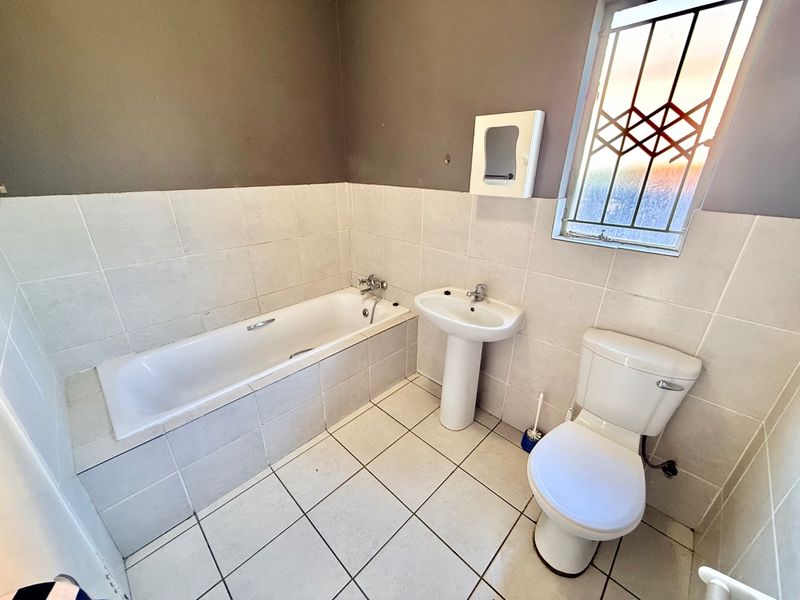 A functional bathroom featuring a bathtub, pedestal basin, and toilet with neutral wall and floor tiling and security bars on the window.