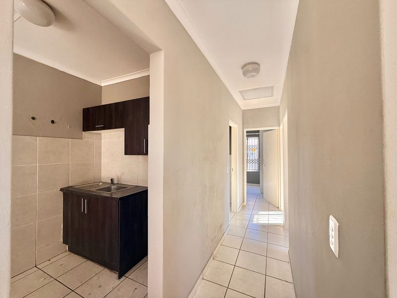 A compact scullery area featuring built-in dark cabinetry, a stainless steel sink, and tiled finishes, leading into a connecting hallway.