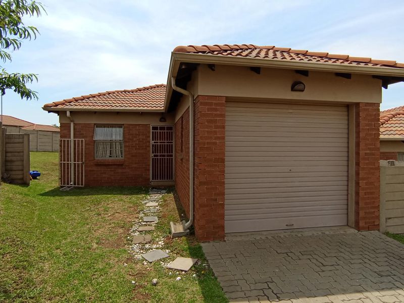 Front view of a single-story brick house featuring a roller garage door, paved driveway, and a small garden area with a precast boundary wall.