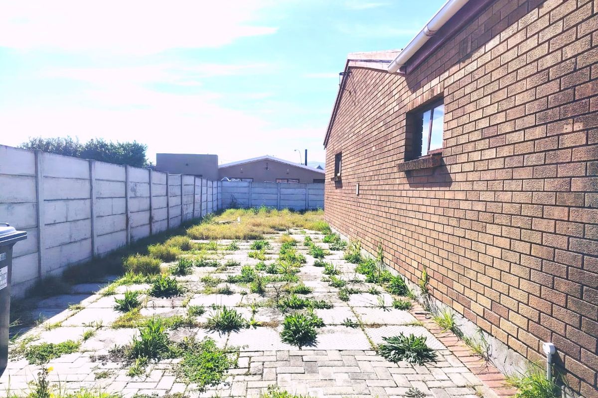 Rear exterior view of the property featuring a face brick wall, paved patio area, and precast concrete boundary wall with significant weed growth.