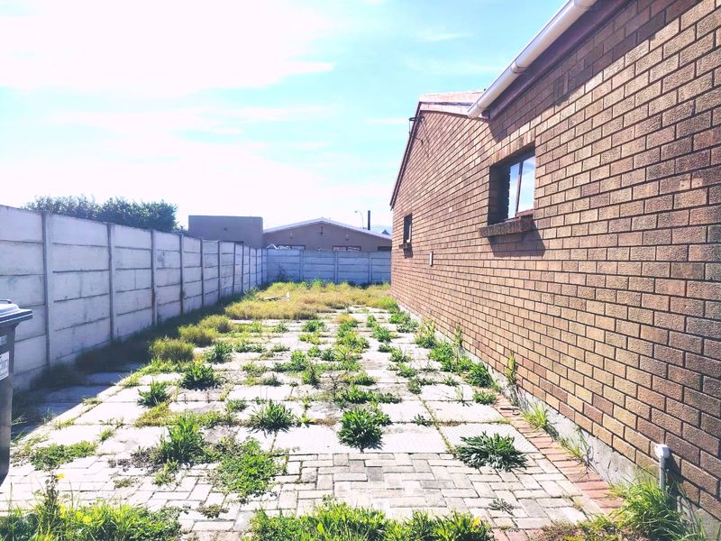 Rear exterior view of the property featuring a face brick wall, paved patio area, and precast concrete boundary wall with significant weed growth.