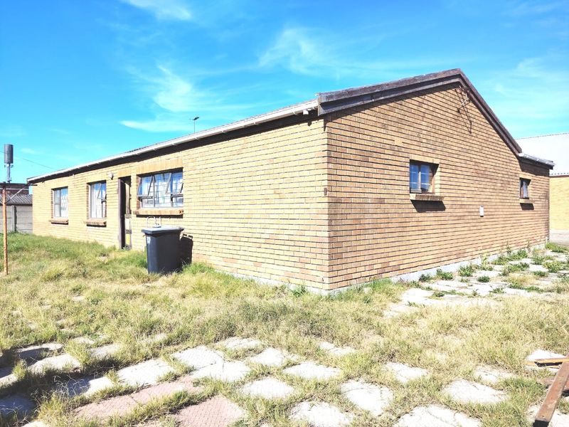 A side and rear view of a face brick house featuring steel frame windows and surrounding paved areas with overgrown grass.