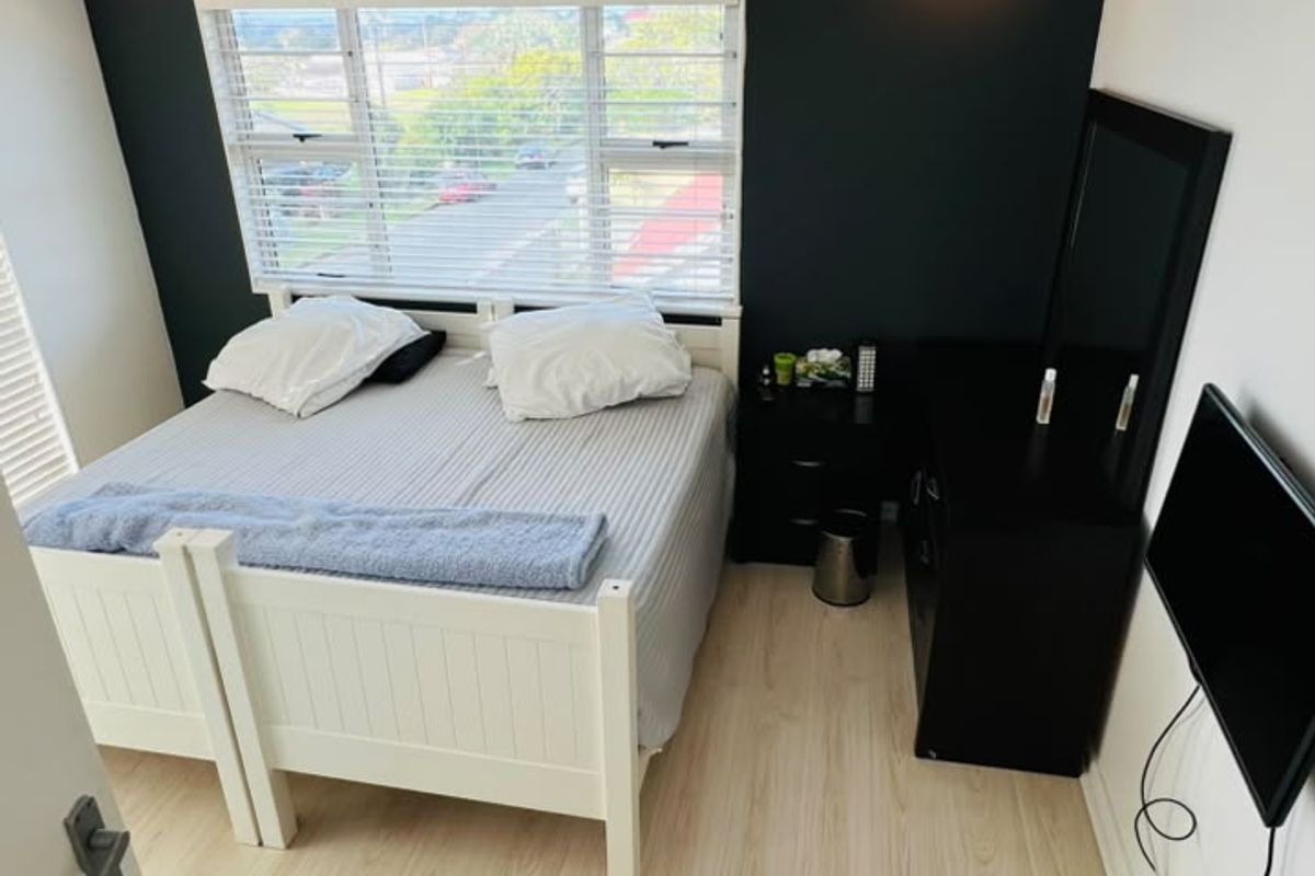 A bedroom featuring light-toned laminate flooring, a dark accent wall, and large windows with fitted venetian blinds.