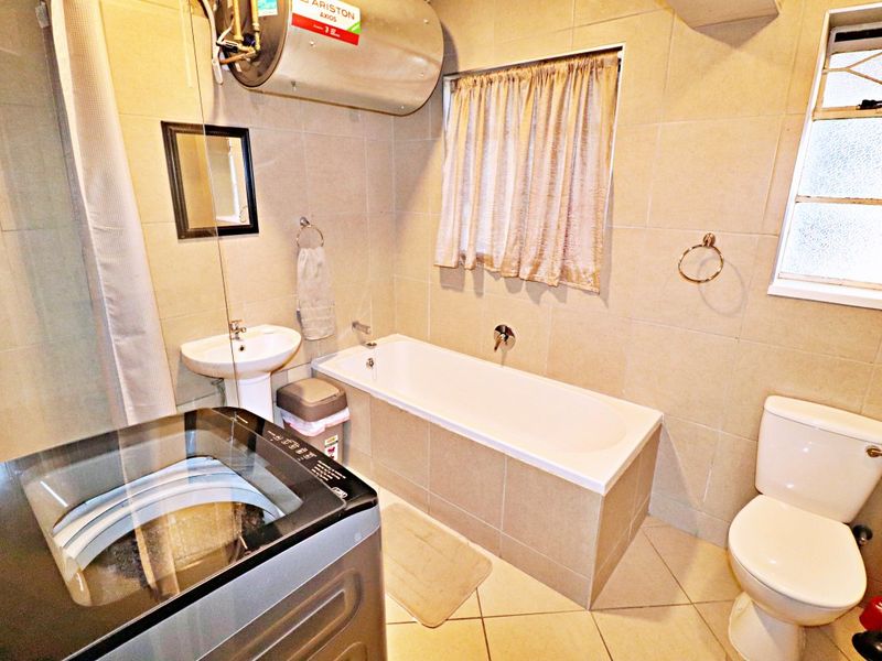A functional bathroom featuring a bathtub, pedestal basin, toilet, and wall-mounted electric water heater, finished with neutral floor and wall tiling.