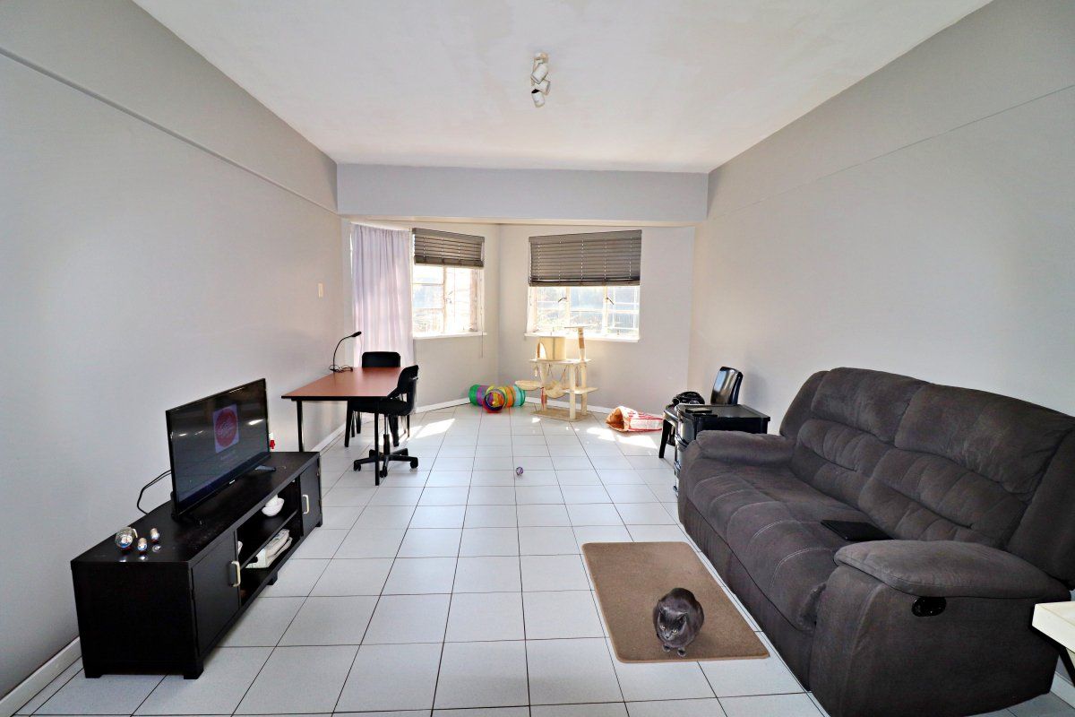 A spacious living area featuring neutral wall tones, tiled flooring, and large windows fitted with venetian blinds, offering a versatile layout for relaxation.