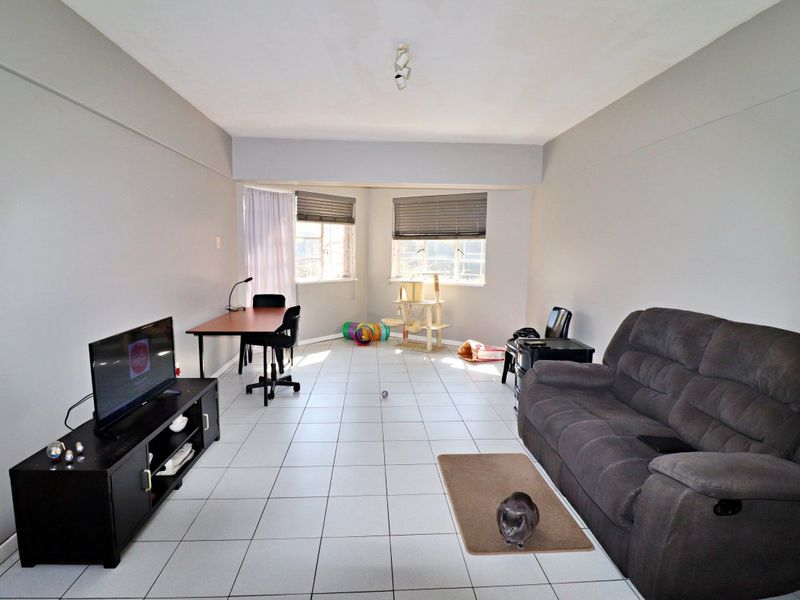 A spacious living area featuring neutral wall tones, tiled flooring, and large windows fitted with venetian blinds, offering a versatile layout for relaxation.