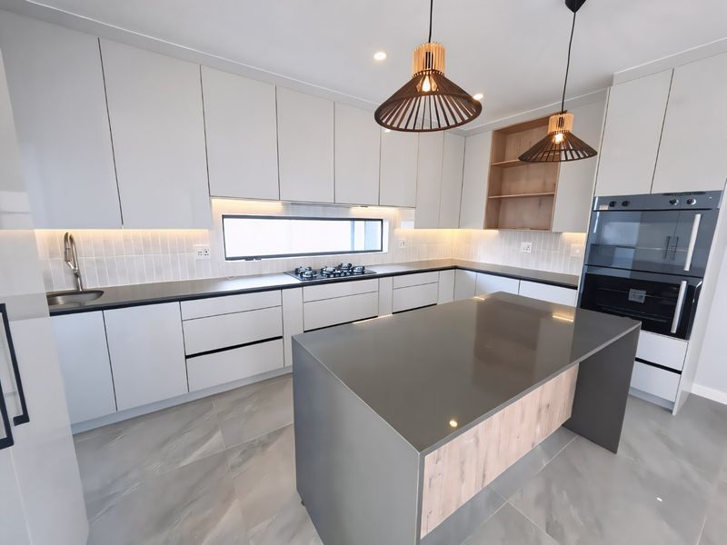 Modern kitchen featuring sleek handleless cabinetry, stone countertops, a central island, built-in double oven, gas hob, and high-quality tiled flooring.