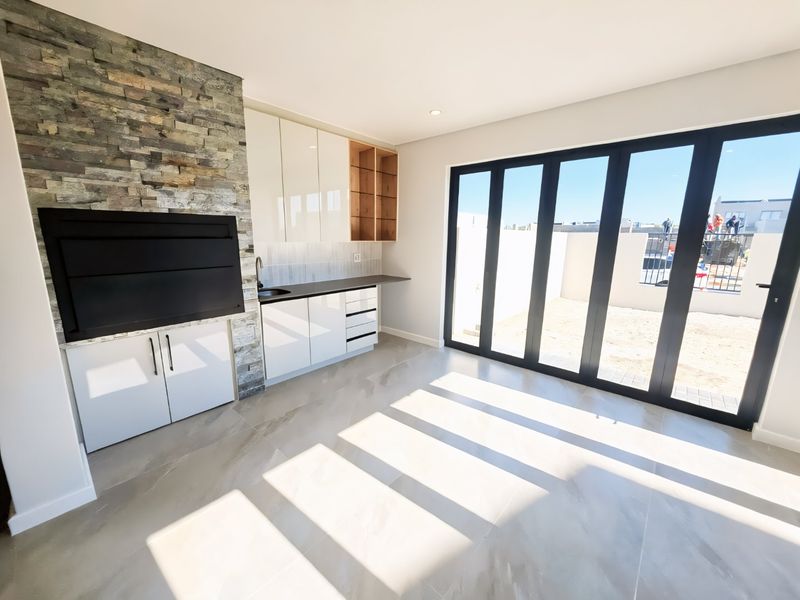 Modern covered patio featuring a built-in braai, stone feature wall, kitchenette with sink, and large stacking doors opening to the rear garden area.