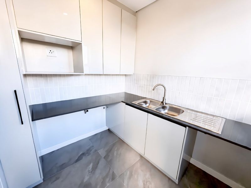 A modern, clean scullery featuring sleek white cabinetry, dark stone countertops, a double stainless steel sink, and contemporary tiled flooring.