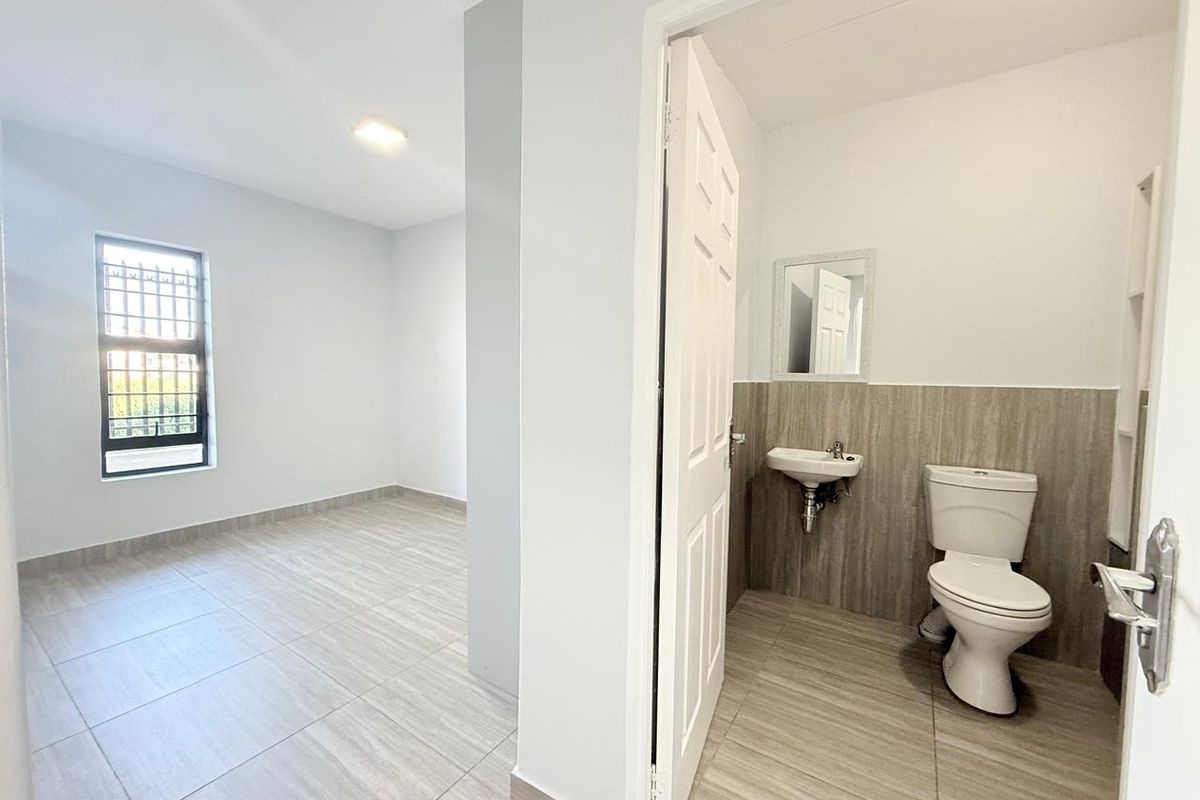 A clean, modern bathroom featuring a wall-mounted basin and toilet, adjacent to an open-plan living area with neutral tiled flooring throughout.