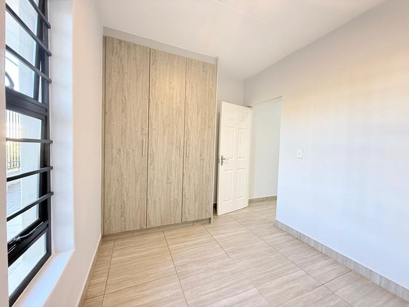 A bright, empty bedroom featuring neutral tiled flooring, a modern built-in wardrobe with wood-grain finish, and a large aluminium-framed window.