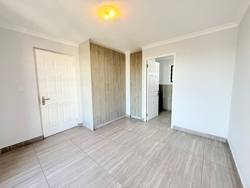 A bright, empty bedroom featuring modern wood-look tiled flooring, built-in cupboards with wood-grain finish, and crisp white walls.