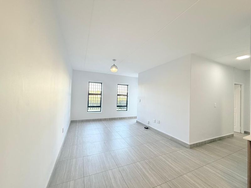 A bright, empty living area featuring neutral tiled flooring, clean white walls, and windows fitted with security bars.
