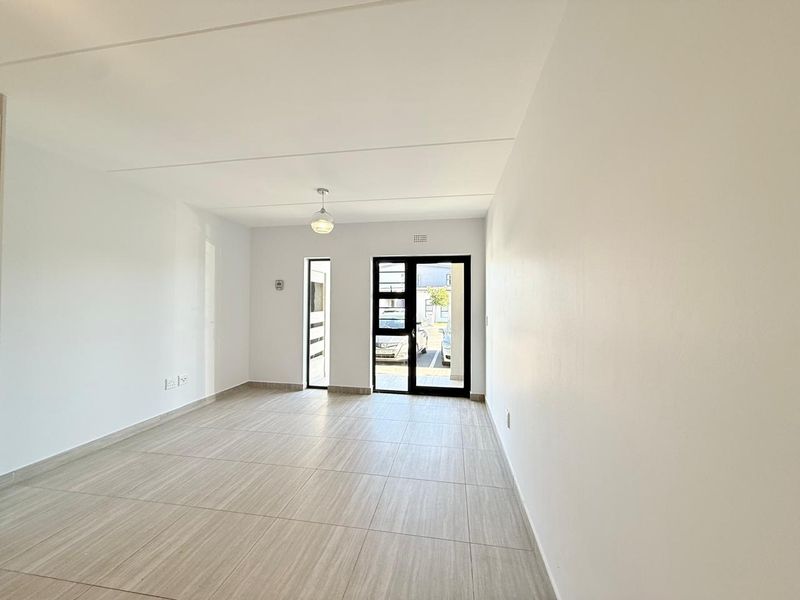 A bright, empty living area featuring neutral tiled flooring, clean white walls, and modern aluminium-framed glass doors leading to the exterior.