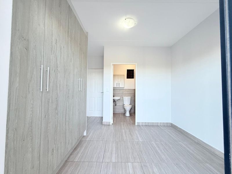 A bright, empty bedroom featuring modern wood-look tiled flooring, extensive built-in cupboards, and direct access to an en-suite bathroom.