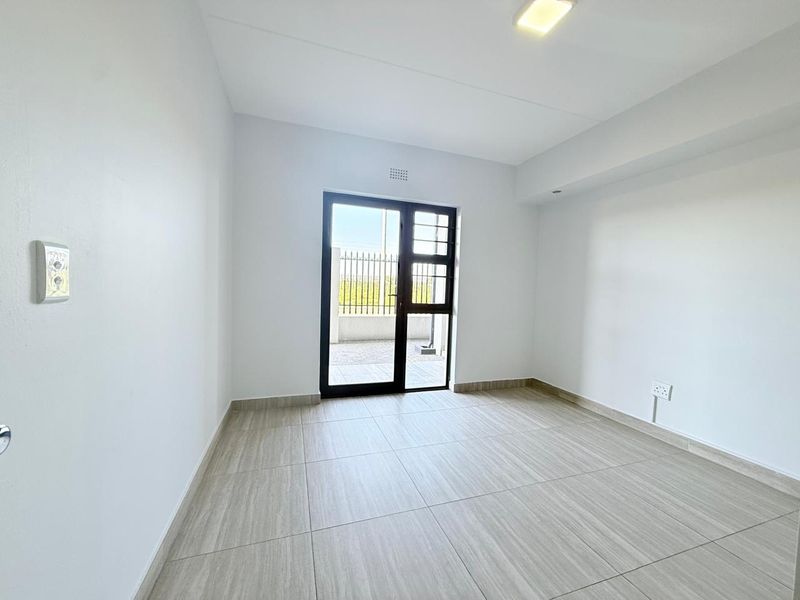A bright, empty living area featuring modern neutral floor tiles, clean white walls, and a glass door providing access to an outdoor space.