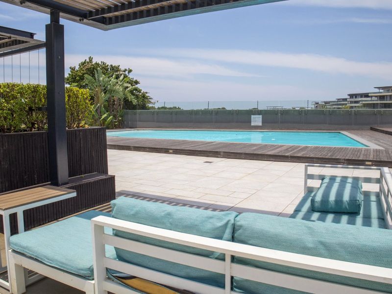 A well-maintained communal pool area featuring timber decking, tiled patio space, a shaded pergola, and a modern glass balustrade overlooking the surroundings.