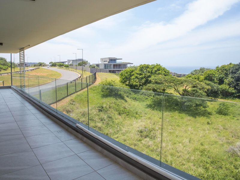 Spacious covered balcony featuring modern tiled flooring and a frameless glass balustrade, offering expansive views of the surrounding landscape and coastal area.