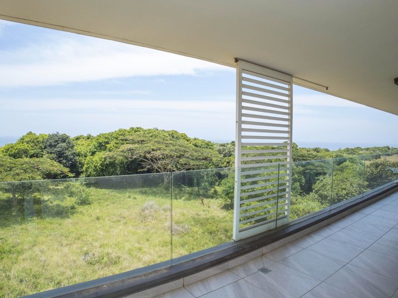 Spacious covered balcony featuring a modern glass balustrade, tiled flooring, and a sliding louvered privacy screen with scenic natural views.