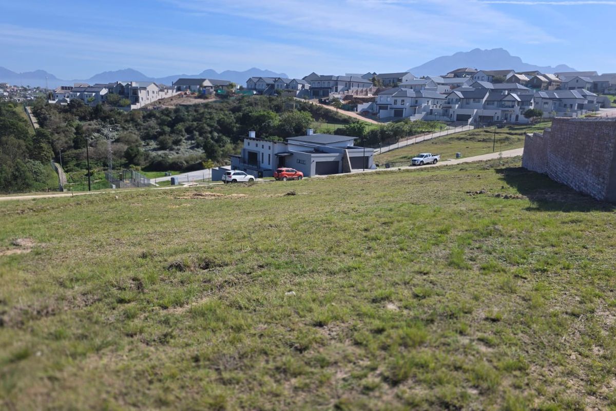 An elevated view of a vacant residential plot featuring a gentle slope and an adjacent retaining wall, overlooking a developed neighborhood.