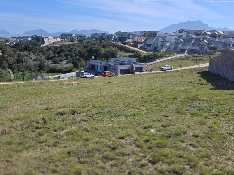 An elevated view of a vacant residential plot featuring a gentle slope and an adjacent retaining wall, overlooking a developed neighborhood.