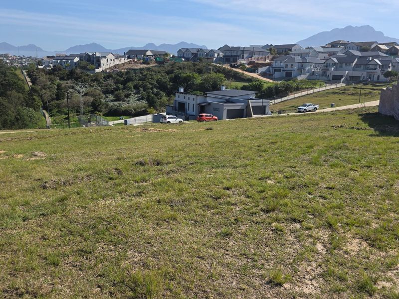 A wide-angle view of a vacant residential plot featuring sloped grassland with surrounding estate housing and mountain views in the distance.