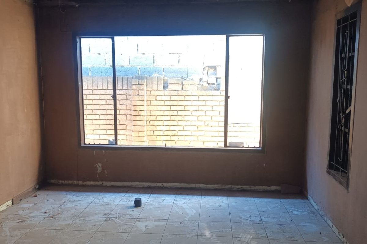 An empty living room space featuring tiled flooring and a large window with security bars, currently requiring wall finishing and general renovation.