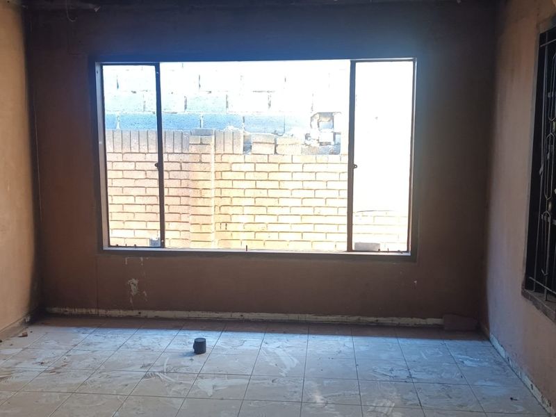 An empty living room space featuring tiled flooring and a large window with security bars, currently requiring wall finishing and general renovation.