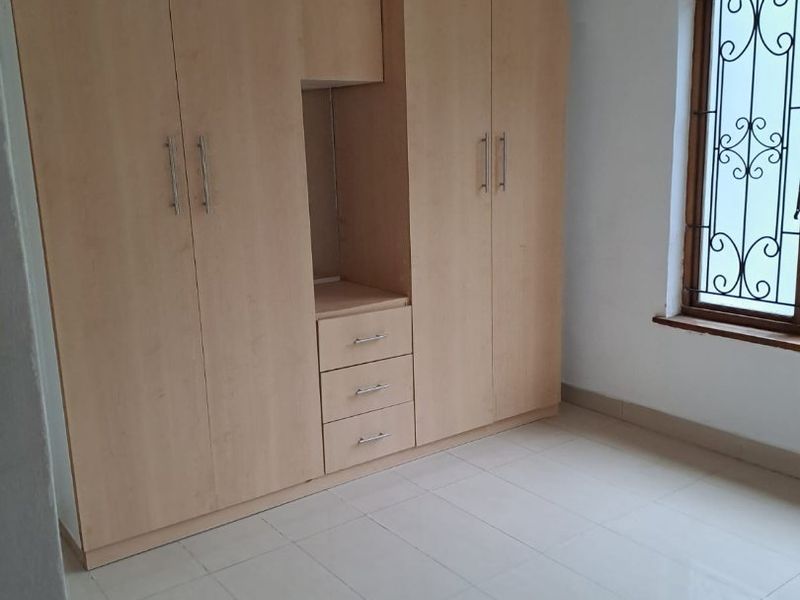 A clean, empty bedroom featuring light wood-finish built-in cupboards, tiled flooring, and a window fitted with decorative security bars.