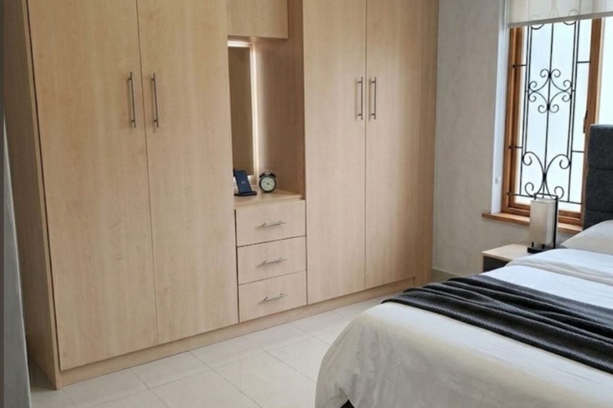 A well-maintained bedroom featuring light wood built-in cupboards, tiled flooring, and a window fitted with security bars and a roller blind.
