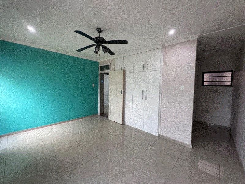 A spacious, empty bedroom featuring neutral tiled flooring, built-in white cupboards, a ceiling fan, and a vibrant teal accent wall.