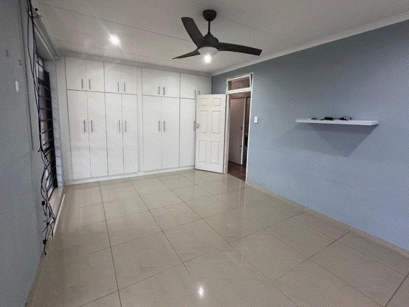 A spacious, empty bedroom featuring tiled flooring, built-in cupboards, a ceiling fan, and a floating wall shelf.