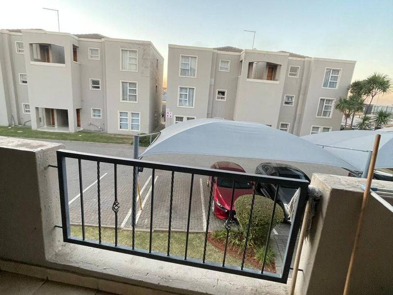 A covered balcony with tiled flooring and a metal balustrade, overlooking the apartment complex parking area and shade-net carports.