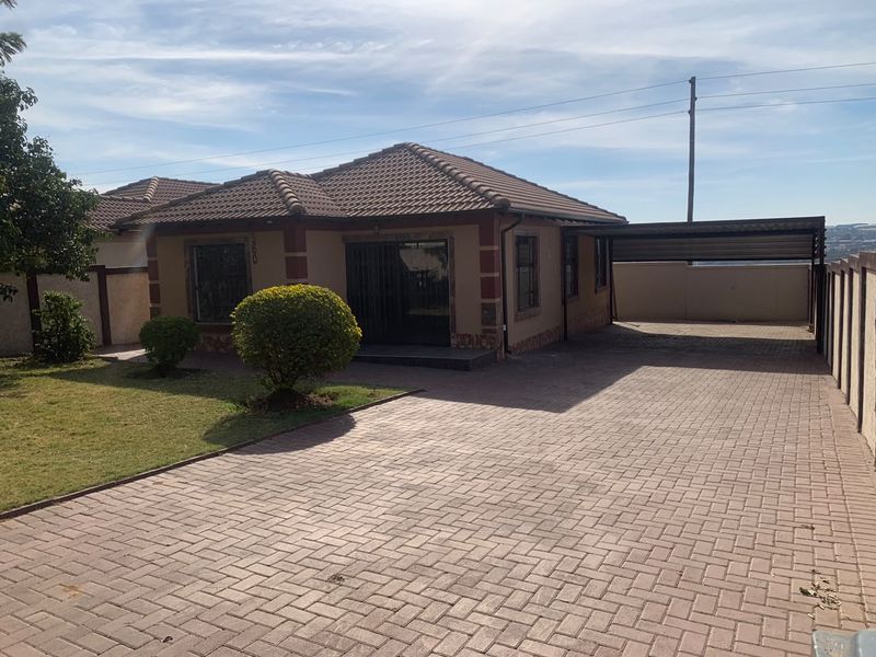 Front view of a single-story house featuring a tiled roof, paved driveway, and a covered carport, set within a secure walled perimeter.