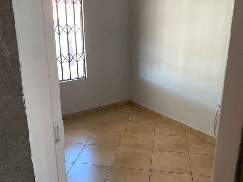 A compact, empty bedroom featuring neutral wall paint, tiled flooring, and a window fitted with security burglar bars.