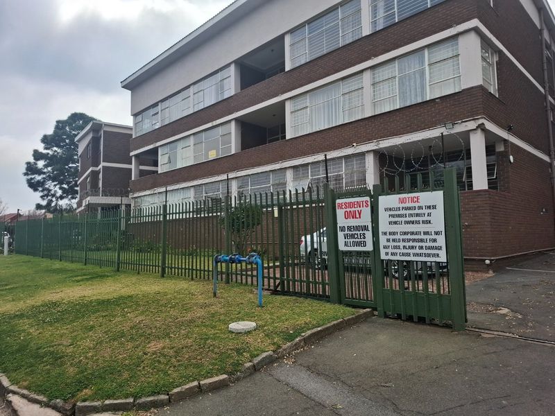 Exterior view of a multi-story brick apartment building featuring secure steel fencing, razor wire, and paved driveway access.