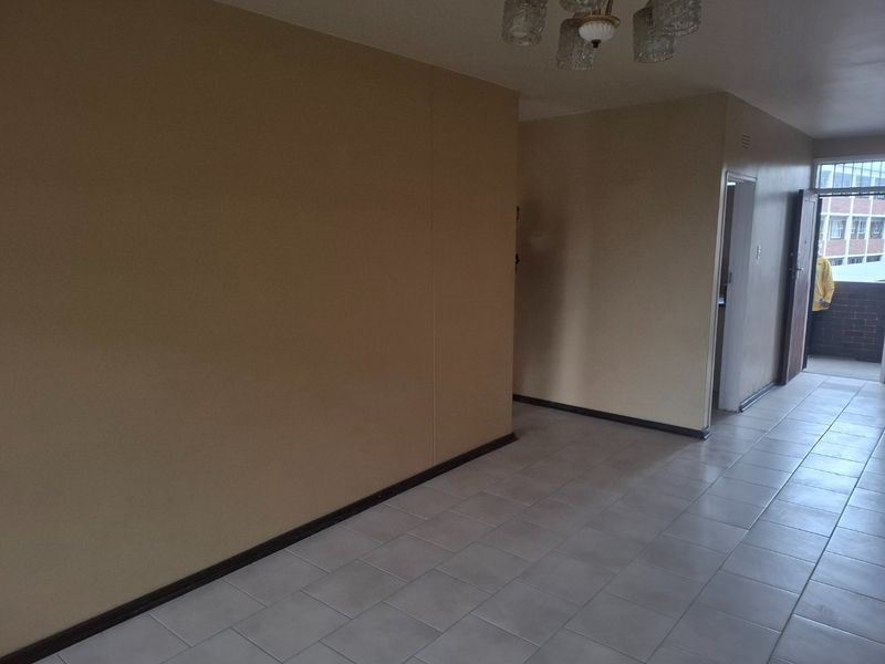 An empty living area featuring neutral wall tones, tiled flooring, and a ceiling light fixture, leading towards an exterior balcony entrance.