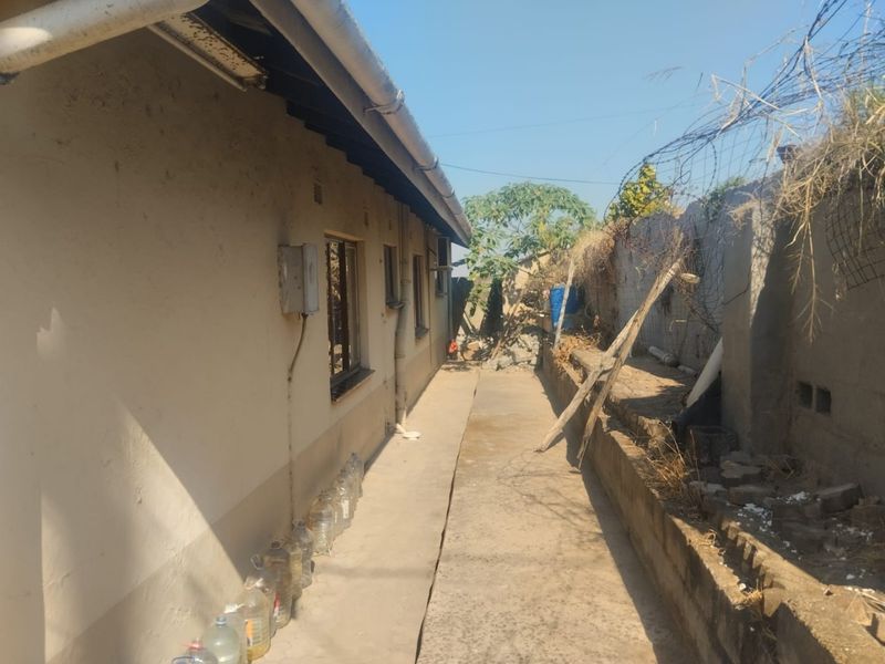 A narrow rear exterior walkway featuring a concrete path, boundary wall, and basic building infrastructure in need of maintenance and repair.