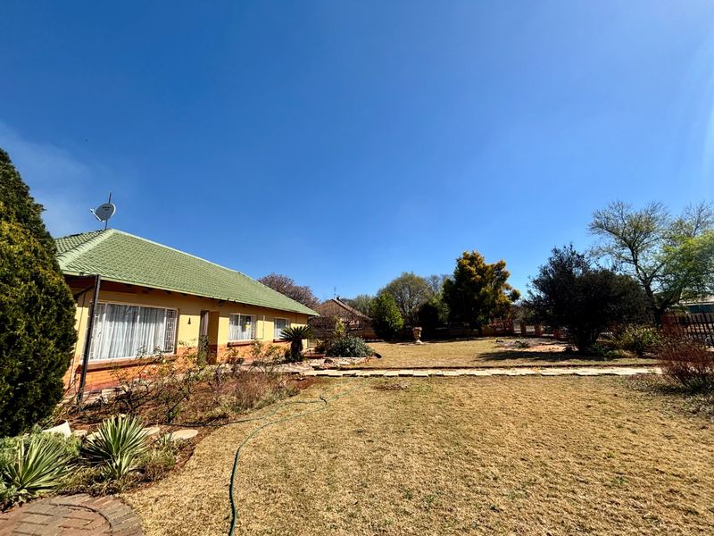 A spacious rear garden featuring a well-maintained brick house with a tiled roof, large windows, and an established lawn area.