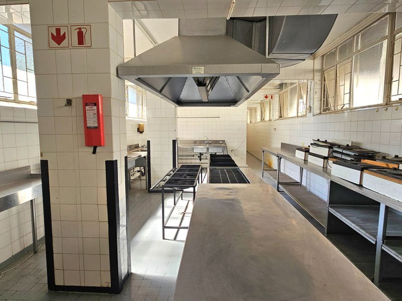 A functional commercial kitchen space featuring stainless steel prep surfaces, a large extractor hood, tiled walls, and essential fire safety equipment.