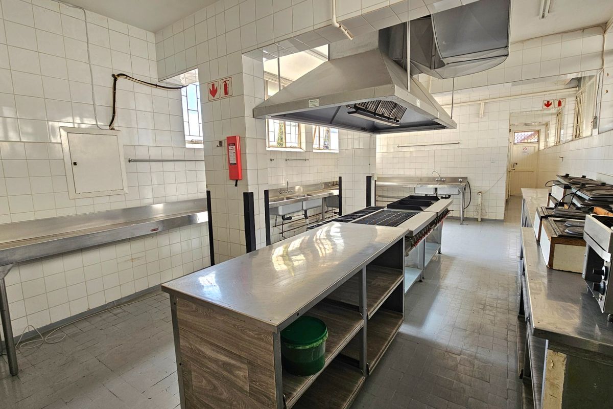 A functional commercial kitchen space featuring stainless steel prep surfaces, a large extractor hood, commercial sinks, and durable tiled wall finishes.