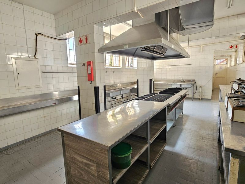 A functional commercial kitchen space featuring stainless steel prep surfaces, a large extractor hood, commercial sinks, and durable tiled wall finishes.