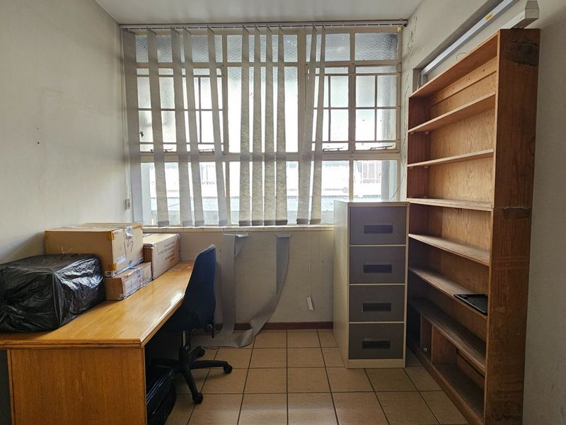A functional office space featuring tiled flooring, built-in shelving, and large windows with vertical blinds, offering a practical layout for a commercial workspace.
