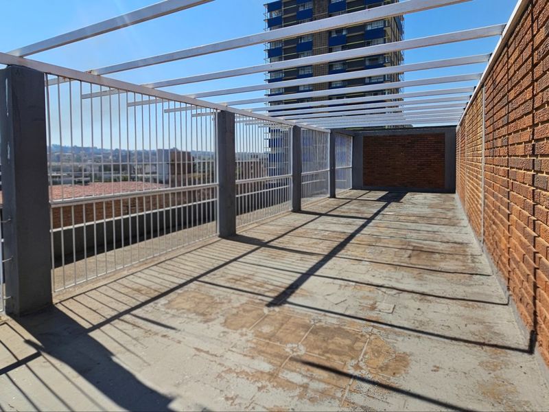 An open-air rooftop balcony featuring exposed brick walls, a metal pergola frame, and security fencing, offering a blank canvas for outdoor commercial use.