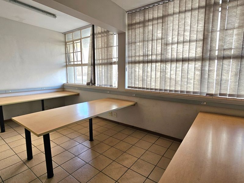A functional office space featuring tiled flooring, built-in desk surfaces, and large windows with vertical blinds, providing ample natural light.