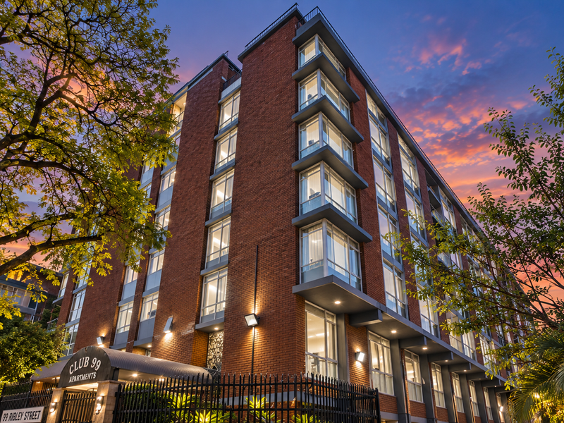 A striking multi-story brick apartment building facade at dusk, featuring large windows, secure perimeter fencing, and a prominent entrance canopy.